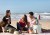 Trzy osoby siedzące na plaży wokół jasnoszarego barku z niebieskim wnętrzem i podniesionym blatem, używanego jako stolik z napojami i przekąskami.