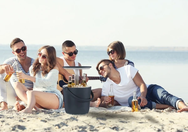 Grupa osób siedząca na plaży wokół ciemnoszarego barku z podniesionym blatem, wypełnionego lodem i butelkami.